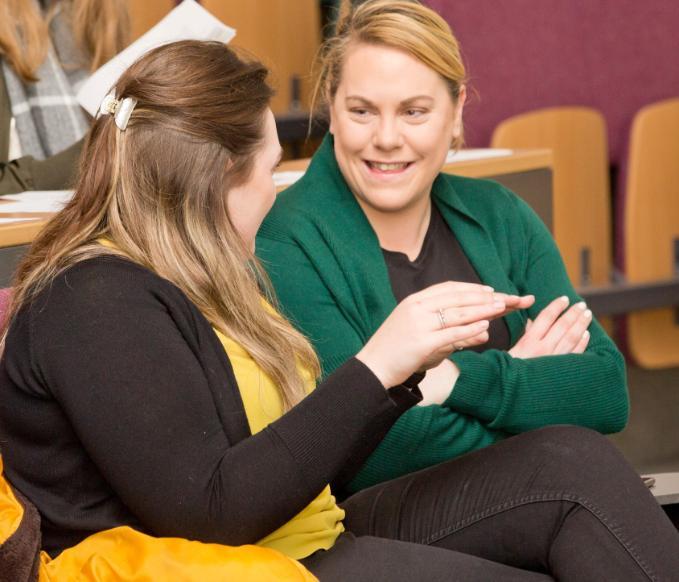 Two female students chatting in a lecture hall while they wait for class to begin