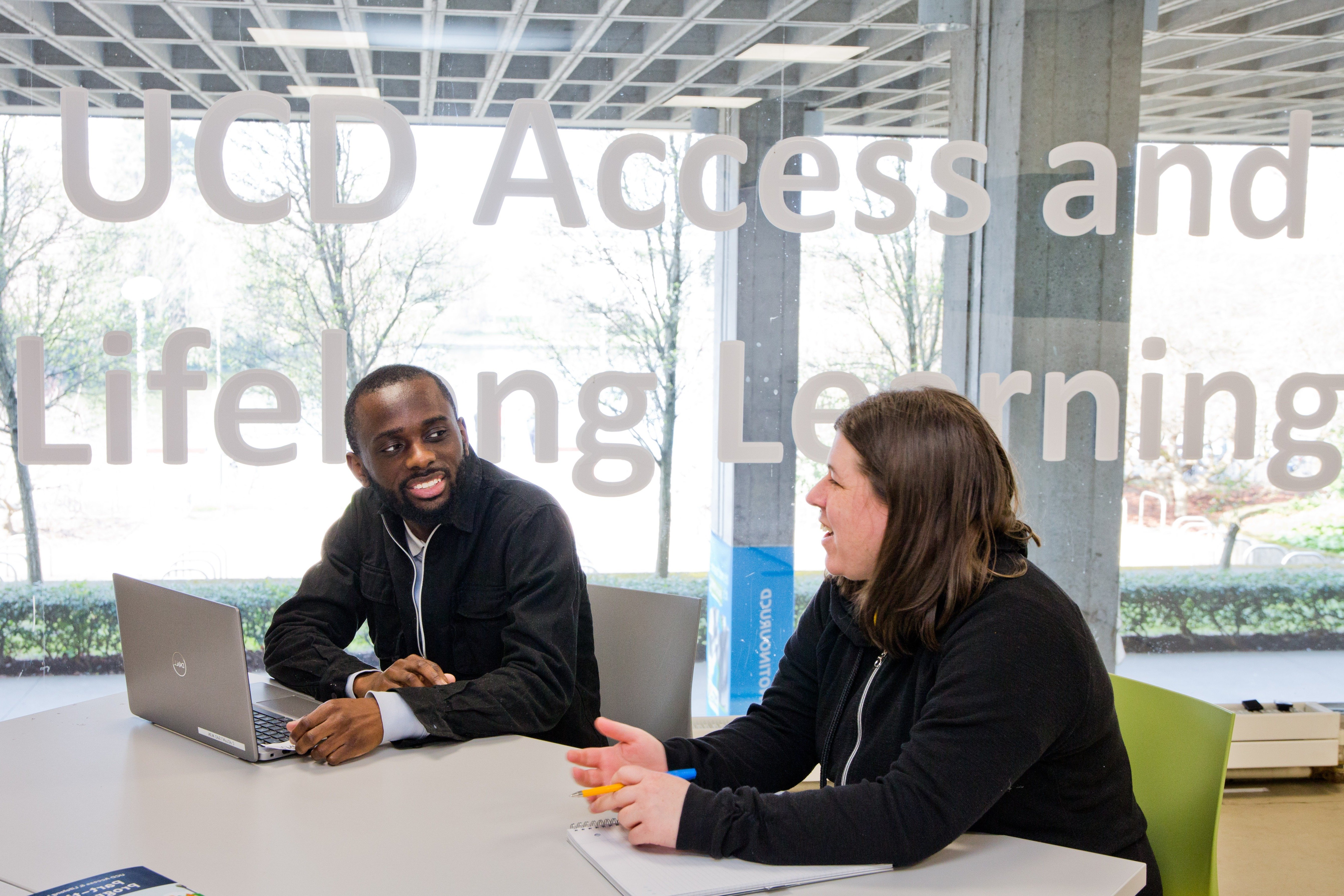 two persons sat at a desk and talking to each other in front of the Access and Lifelong Learning sign