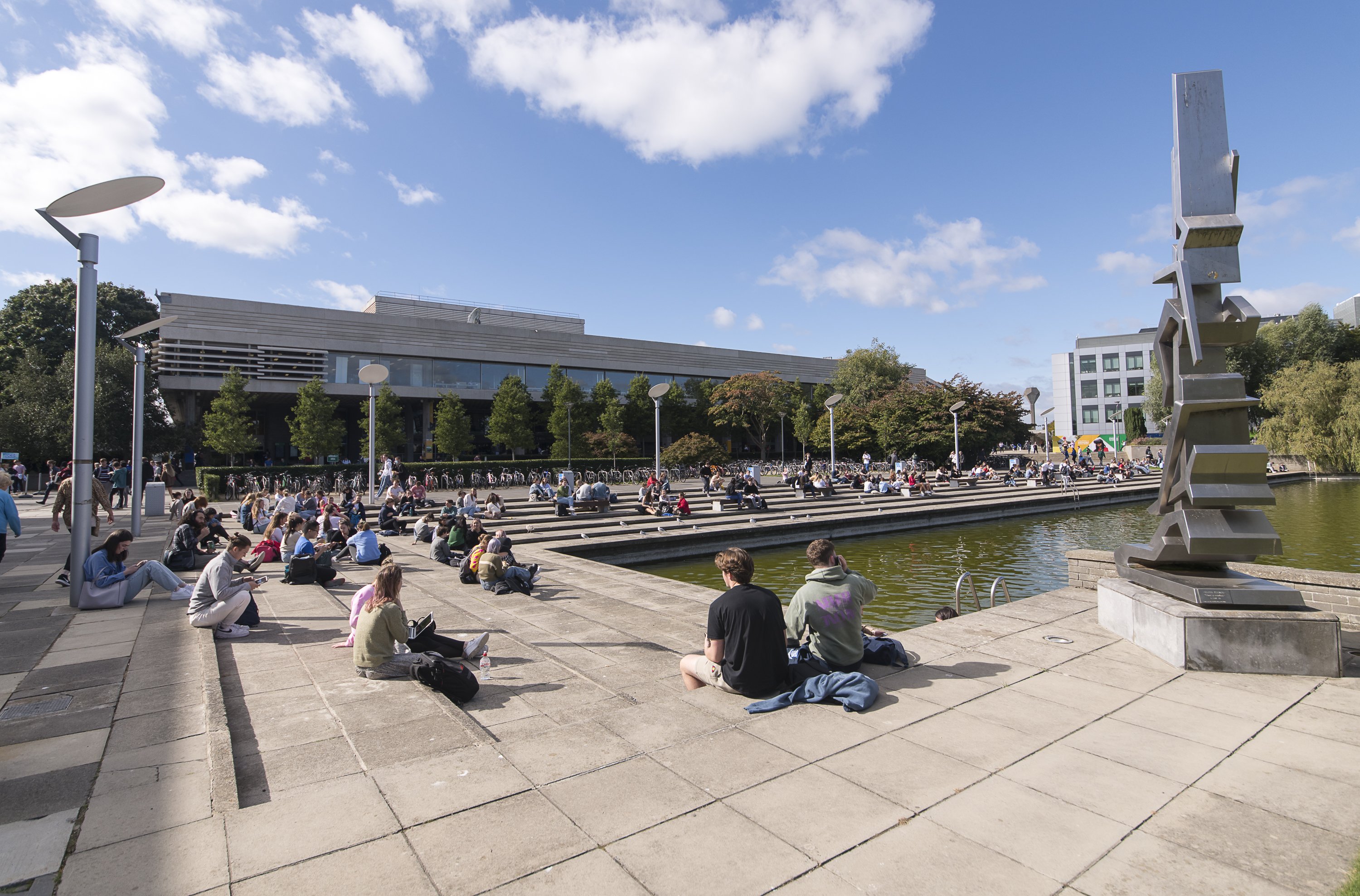 Freshers week - large amount of students enjoying the fine weather sitting around the Belfield Lake, with the James Joyce Library and sculpture