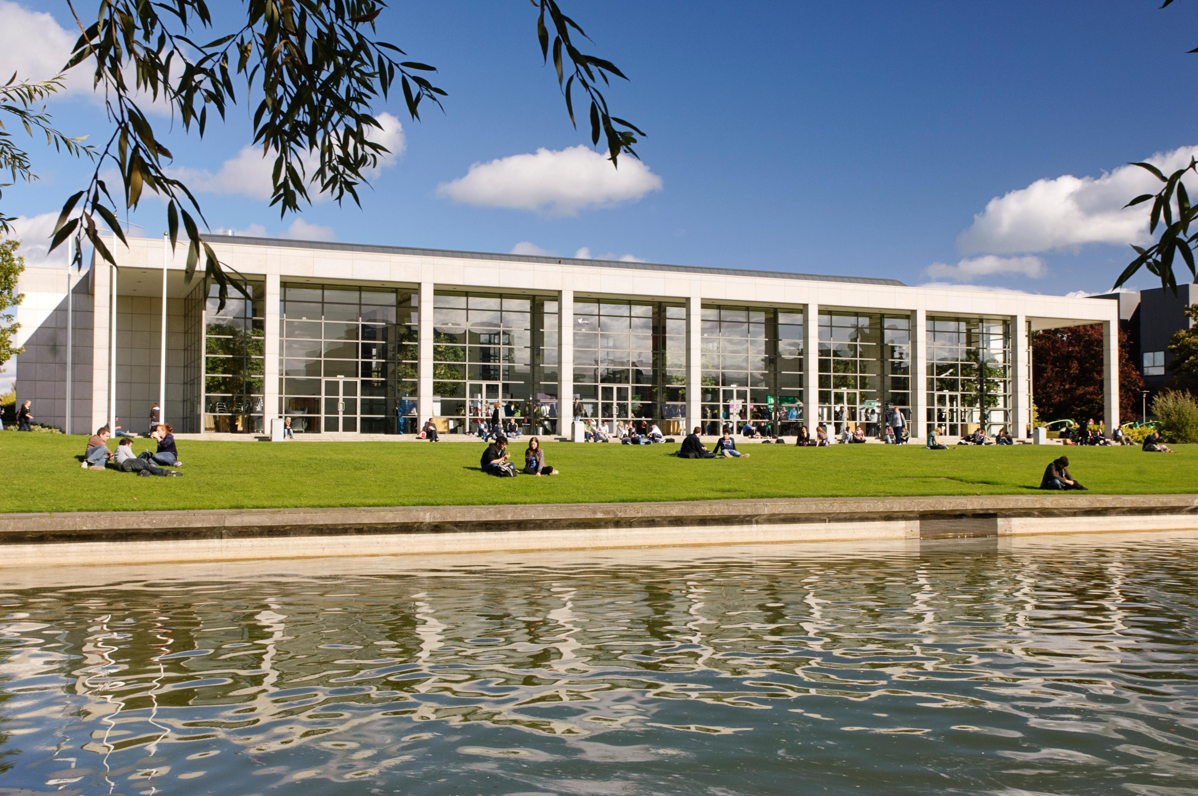 A glass-fronted building with pillars in front of a body of water on a sunny day