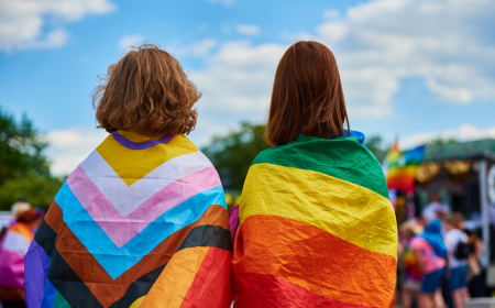 Two people with long hair wearing a progress flag and a pride flag stand next to each other looking at a blue sky with white clouds