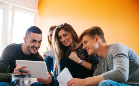 A group of two male and two female students are gathered around an ipad, smiling