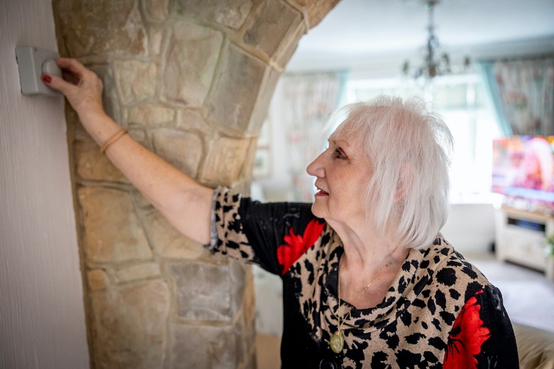 An elderly woman in her home, reaching out to turn a thermostat down