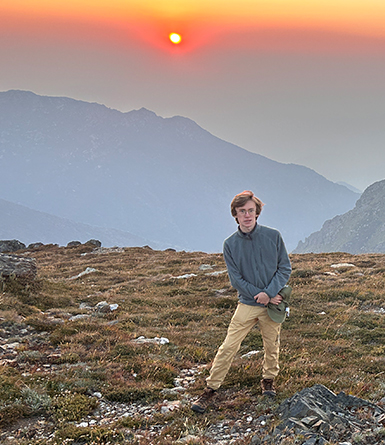 A young person stands on a rocky mountain plateau at sunset, wearing khaki pants and a gray sweater.