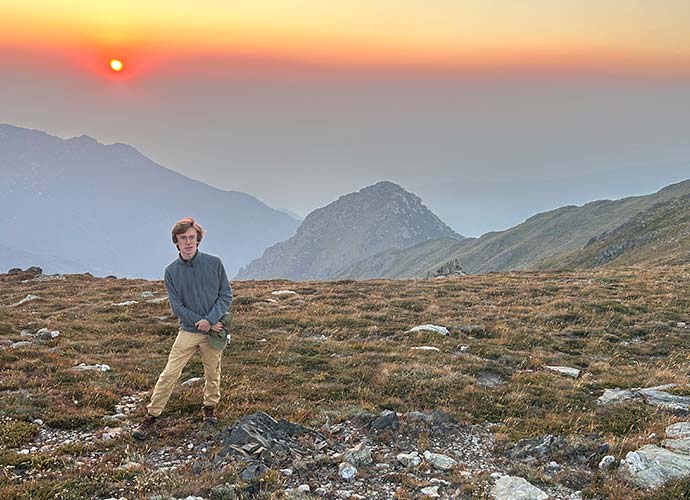 A young person stands on a rocky mountain plateau at sunset, wearing khaki pants and a gray sweater.