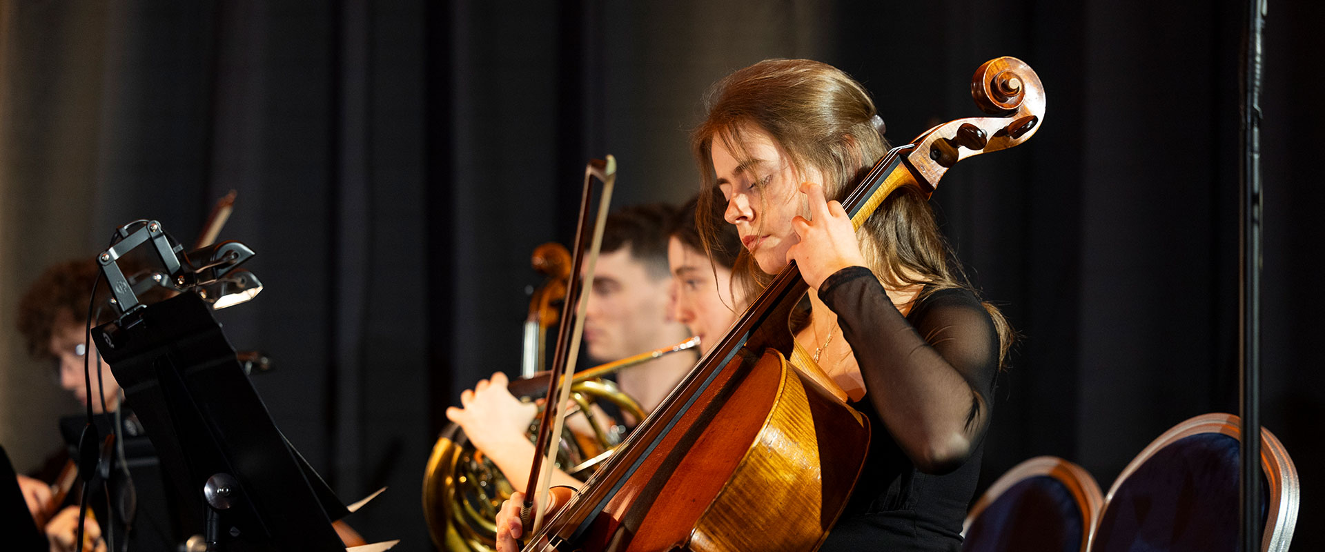A group of students playing in an orchestra. The young woman in the foreground is playing a cello.