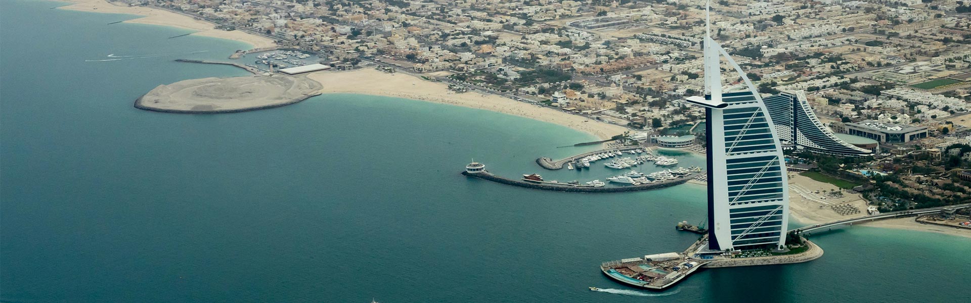 Aerial view of Dubai’s coastline with the sail-shaped Burj Al Arab hotel, marina, and sandy beaches along turquoise water.