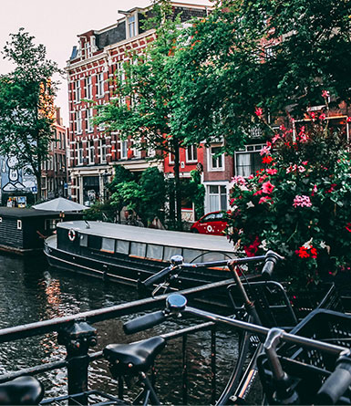 Bicycles parked along a canal in Amsterdam, with a houseboat, red brick buildings, and flowers in the foreground.