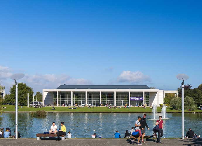 UCD campus lake with people relaxing and the modern university building in the background.