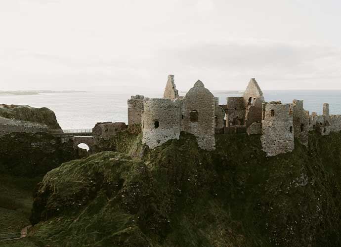 Ruins of an ancient stone castle perched on a cliffside overlooking the sea, with rugged green landscape in the foreground.