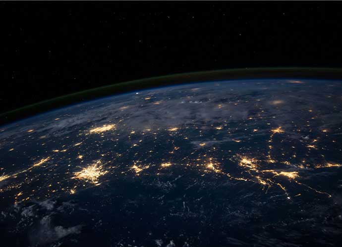 View of Earth from space at night, showing illuminated cities and networks of lights across continents beneath a dark starry sky.