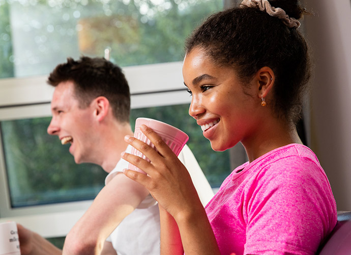 Young woman in a pink t-shirt drinks from a pink cup. A man in a white shirt can be seen laughing in the background.