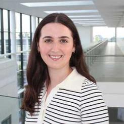 Portrait of a woman with long dark hair, wearing a white collared top with black stripes, smiling in a bright modern building with large windows.