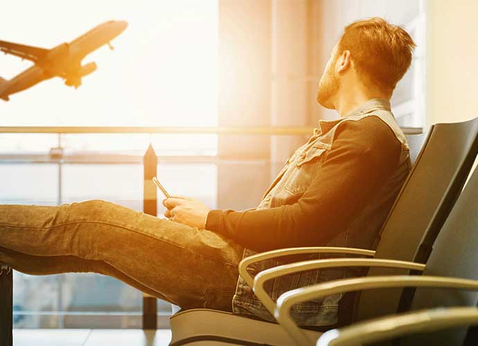 A man sits with his legs on a bench at an airport. A plane ascends in the background.