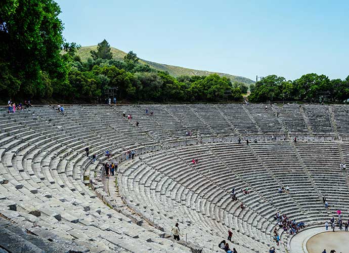Ancient stone amphitheatre with curved seating, surrounded by trees and hills under a clear blue sky.