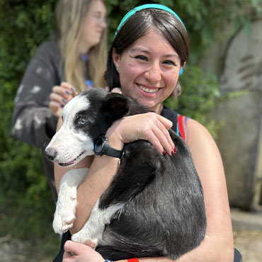 A smiling student holding a black and white dog in their arms, with greenery and another person blurred in the background.