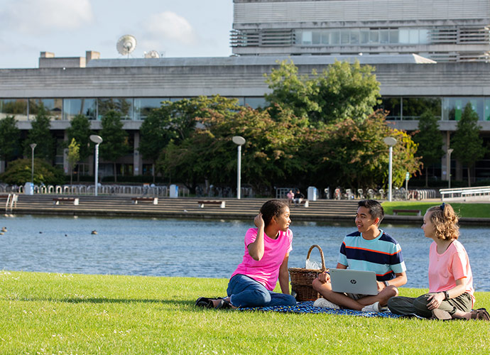 Three students sitting on the grass by a lake on campus, talking and smiling with a laptop and picnic basket beside them.