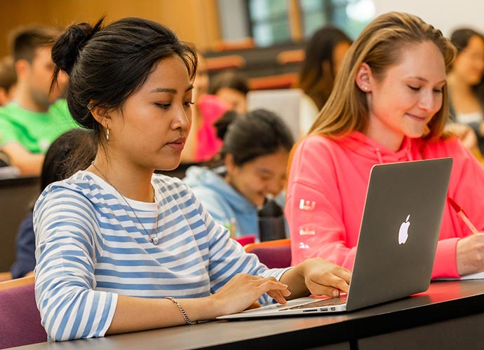 Two students sit in a lecture hall listening intently and using a laptop.