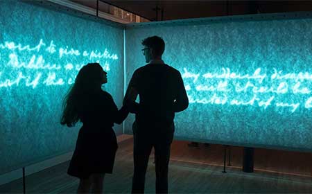 Two people stand close together, looking at glowing blue illuminated text projected onto textured walls in a dark exhibition space.