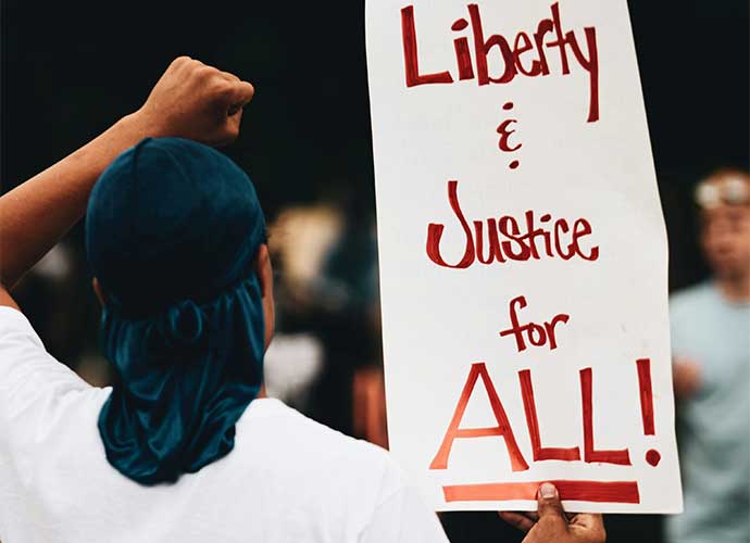Person at a protest holding a sign that reads “Liberty & Justice for ALL!” with a raised fist gesture.