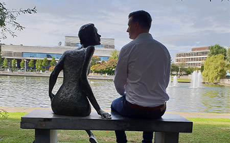 Man sits beside a statue alongside a lake.