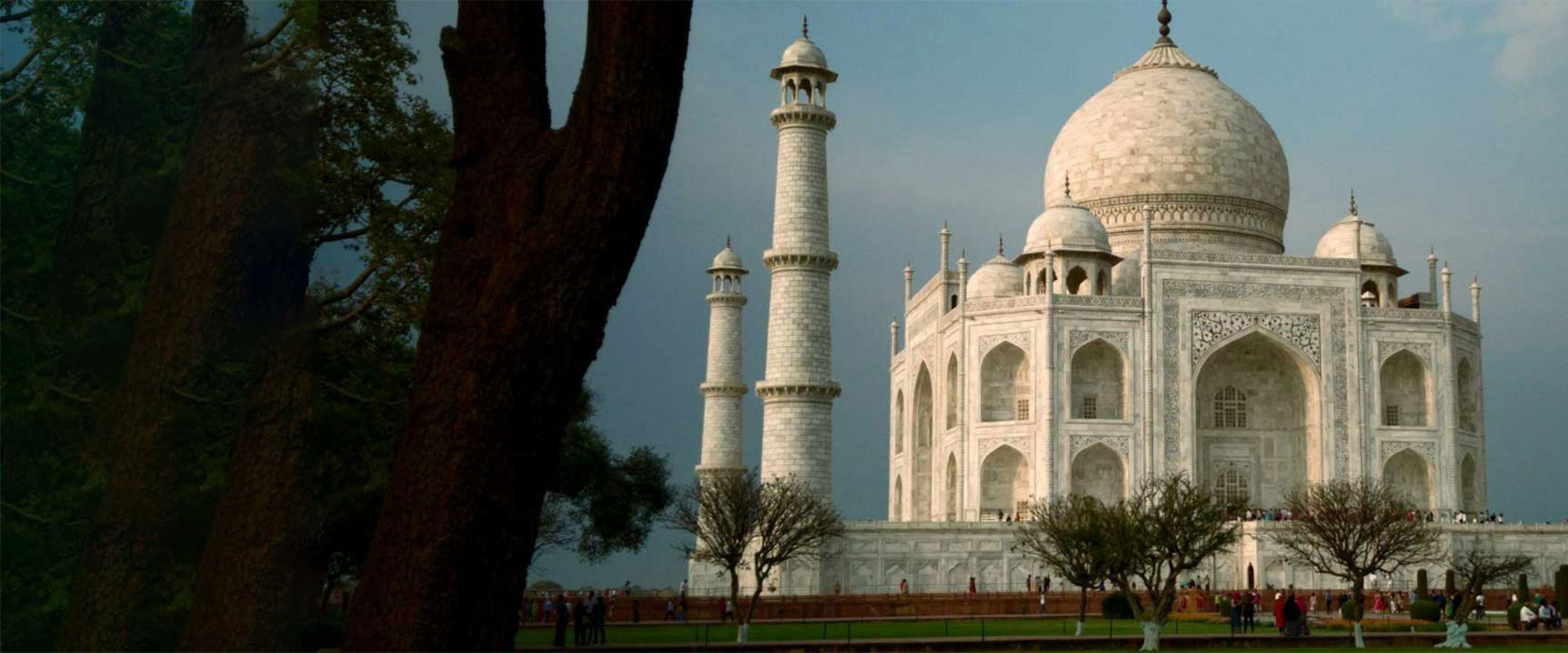 The Taj Mahal in Agra, India, with its white marble dome and minarets, set against a cloudy sky and framed by trees in the foreground, with visitors walking around the base.