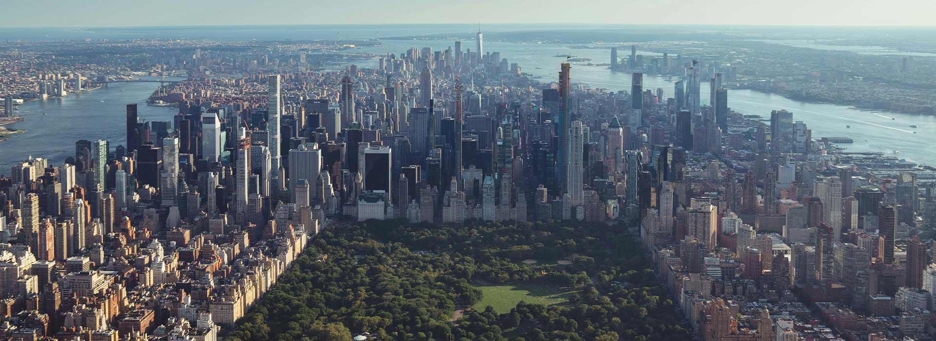 A wide aerial view of Manhattan showing Central Park in the foreground, surrounded by dense high-rise buildings, with the Hudson and East Rivers visible on either side and the city skyline stretching into the distance.