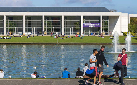Large glass building in front of a man-made lake. Students mill about in the foreground.