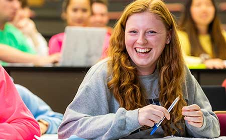 Student smiling in a classroom, holding a pen, with classmates in the background.