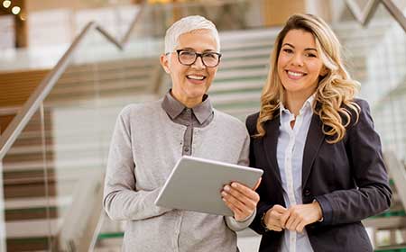 Two professional women standing in an office setting, smiling, one holding a tablet.