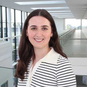 Portrait of a woman with long dark hair, wearing a white collared top with black stripes, smiling in a bright modern building with large windows.