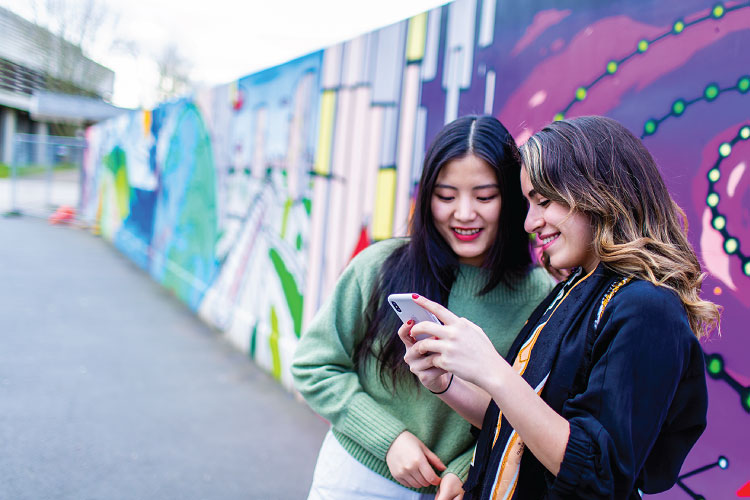 Two female students look at a phone display and smile. There is a colouful wall mural behind them with a science theme on the UCD campus.
