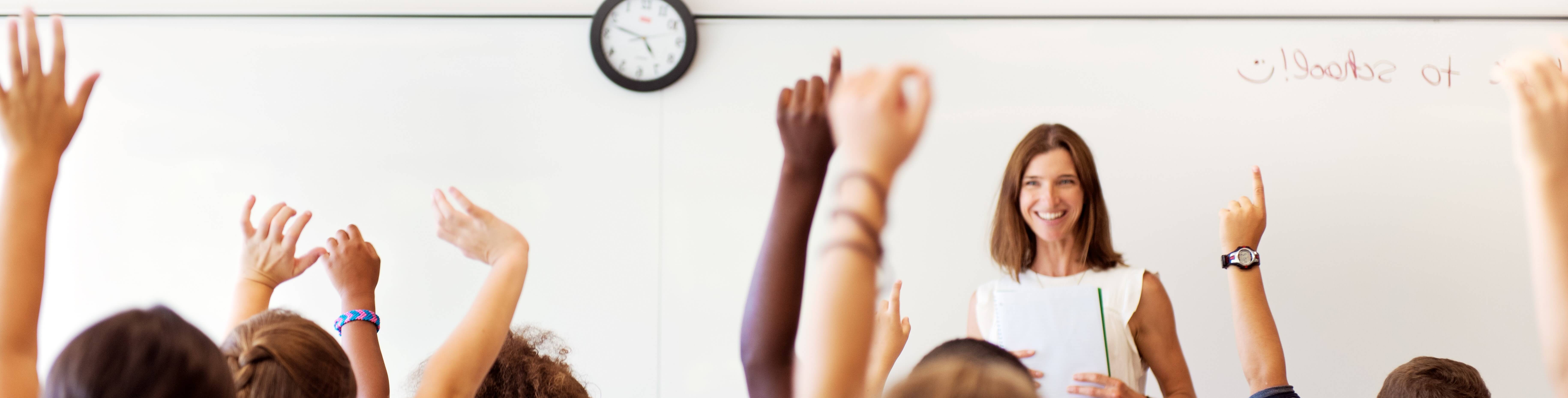 Education course teacher in front of a whiteboard with students hands raised in the foreground