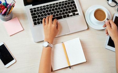 Hand on laptop keyboard with notebook, pens and coffee