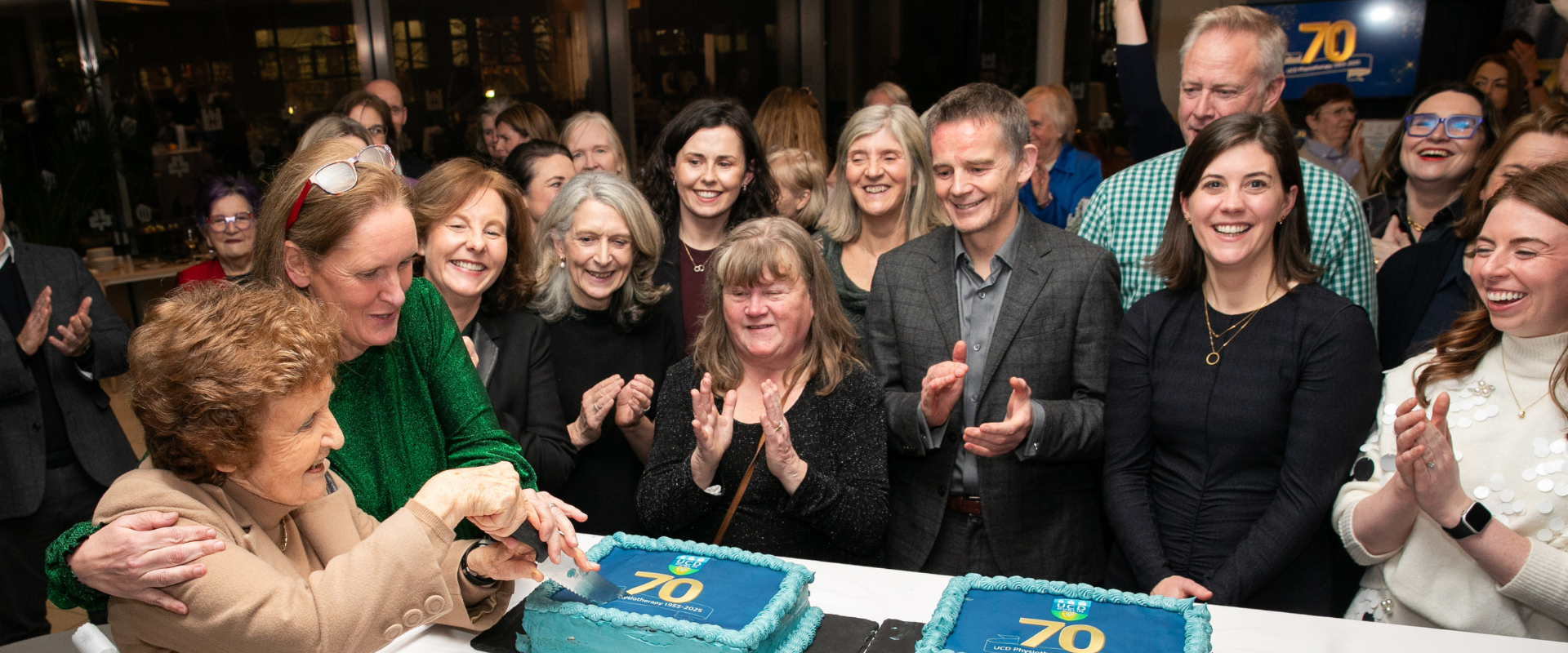 Woman cutting a cake surrounded by a group of people at the UCD Physiotherapy 70th anniversary celebration