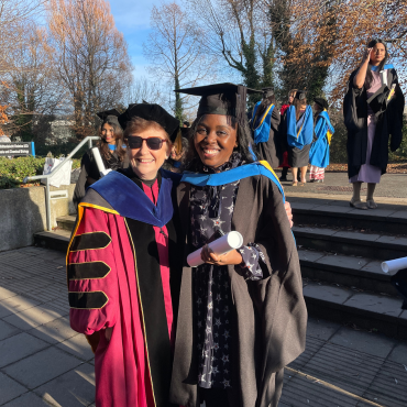 A UCD graduate and a UCD academic in caps and gowns outside on a sunny graduation day.