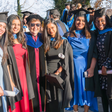UCD graduates and UCD academic in caps and gowns outside on a sunny graduation day.