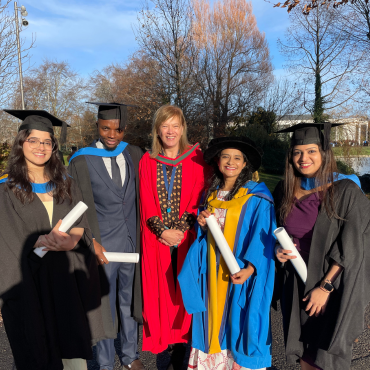 UCD graduates and UCD academic in caps and gowns outside on a sunny graduation day.