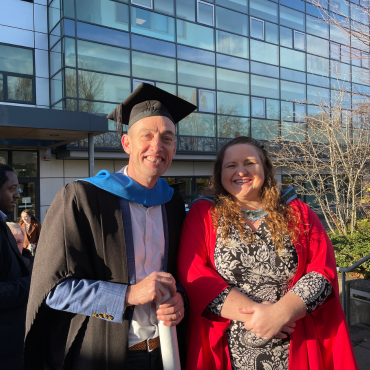 A UCD graduate and an academic in caps and gowns outside on a sunny graduation day.