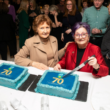 Woman cutting a cake surrounded by a group of people at the UCD Physiotherapy 70th anniversary celebration
