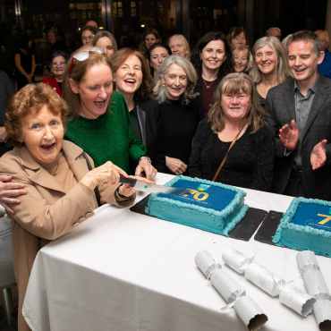 Woman cutting a cake surrounded by a group of people at the UCD Physiotherapy 70th anniversary celebration