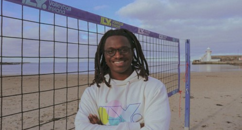 Man with dreadlocks on a beach beside a volleyball net smiling to camera