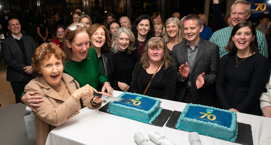 A woman cutting a cake surrounded by a group of people at the UCD Physiotherapy 70th anniversary celebration