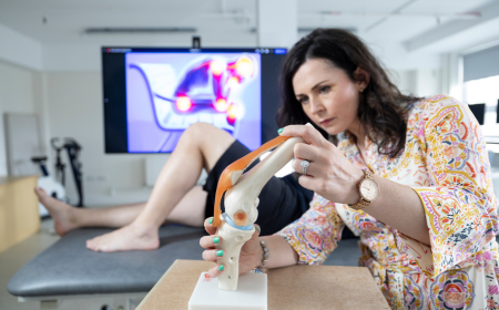 Female physiotherapist examining a model knee joint with a patient's legs and TV monitor visible in background