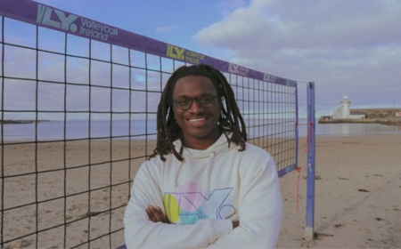 Man with dreadlocks on a beach beside a volleyball net smiling to camera