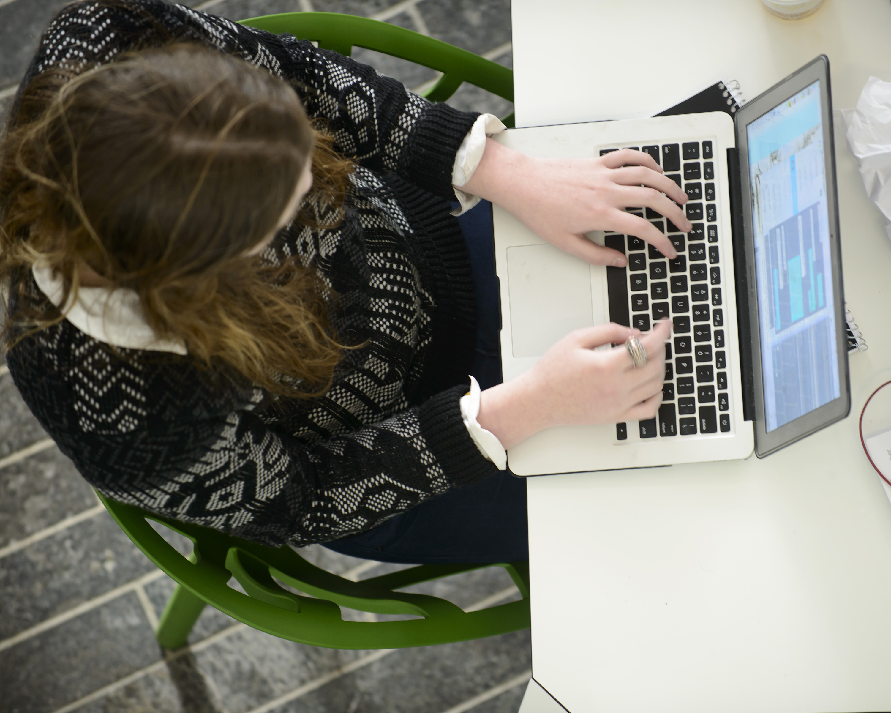 Taken from above this photos shows a woman sitting in a chair, at a desk, typing on a laptop.