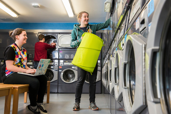 A woman loads her dirty laundry into a washing machine with her clothes basket in her hand