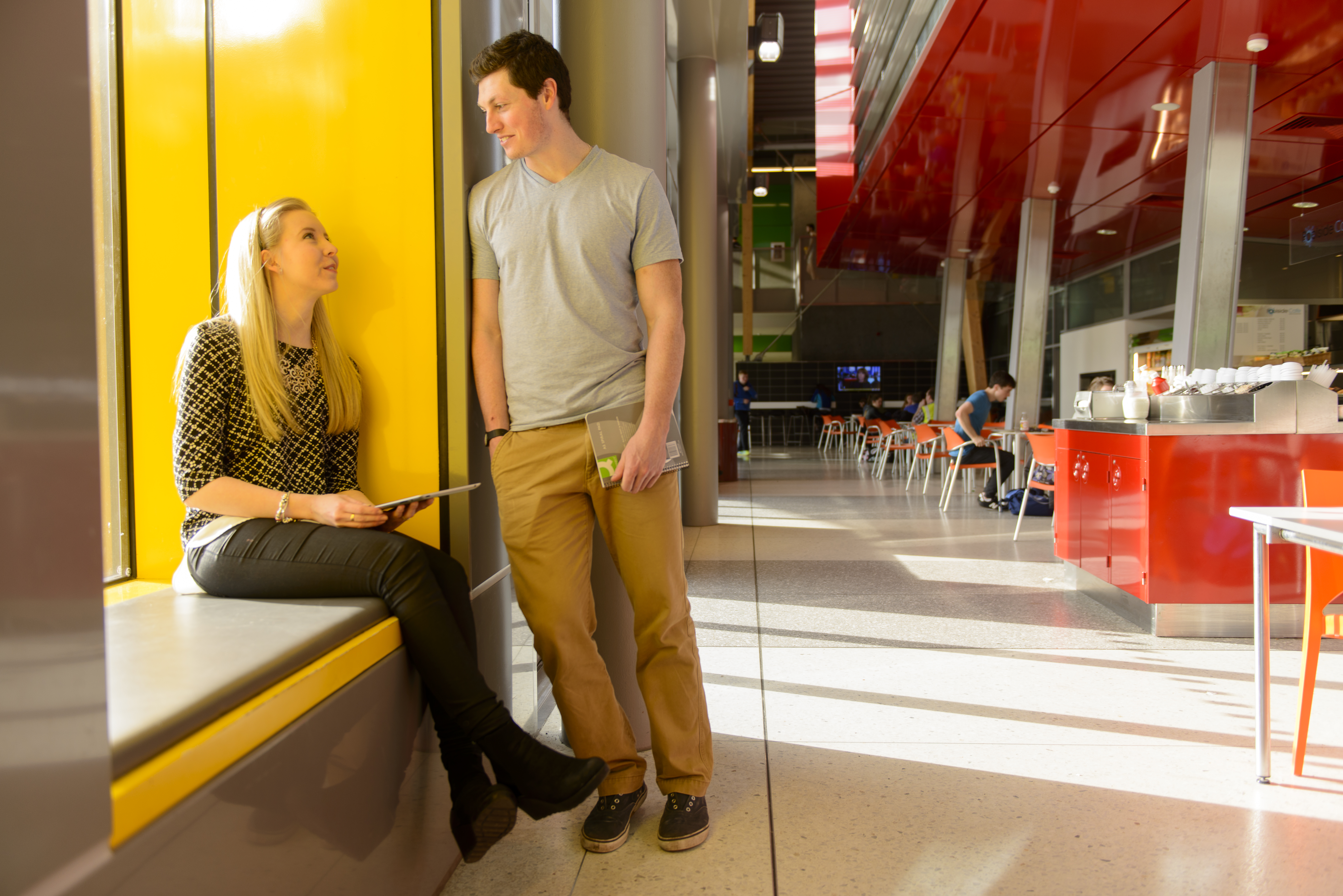 The image shows a female student sitting in a window seat, looking up at a male student with whom she is engaged in conversation and who is leaning on the wall next to her.