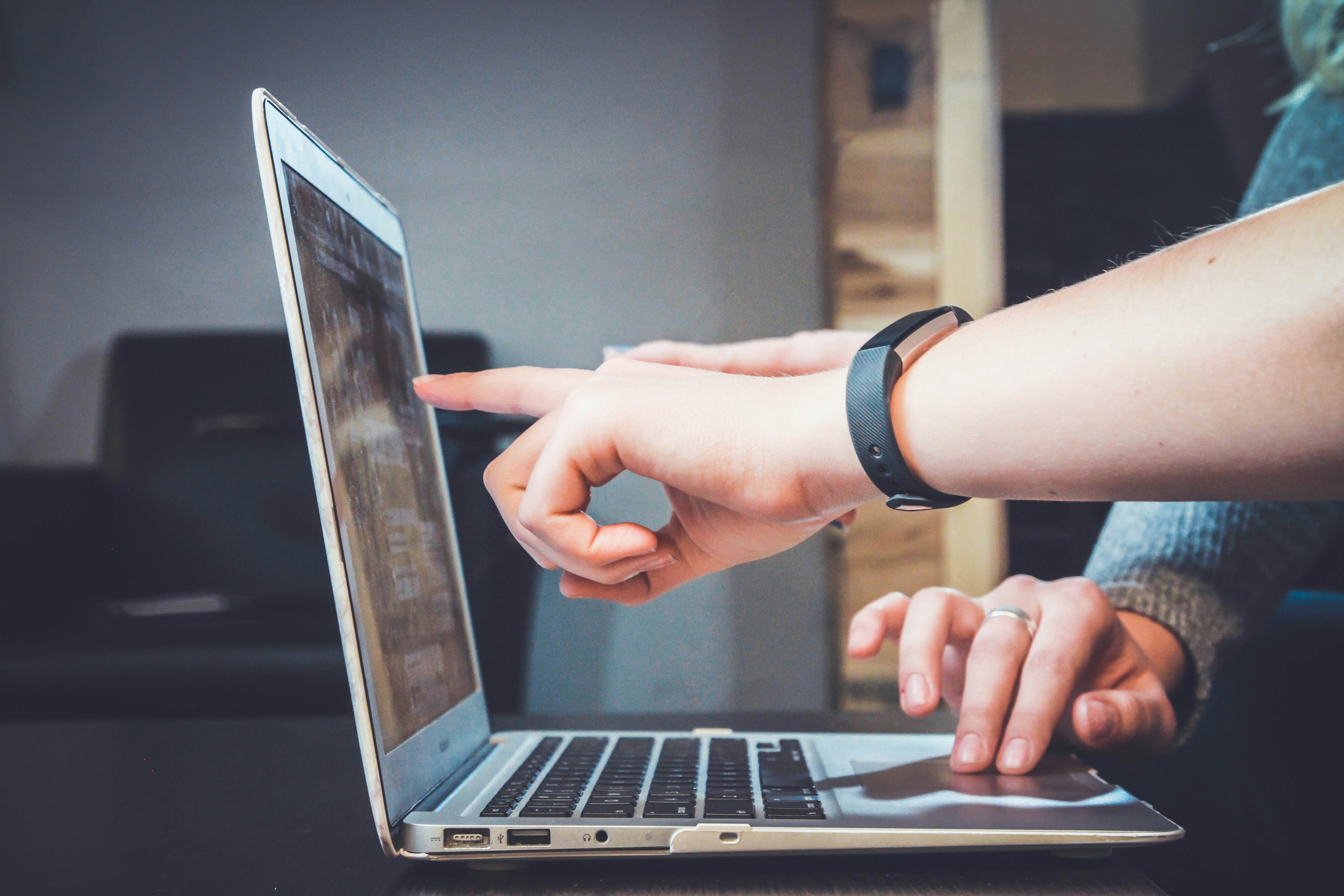 Image shows the hand of a person navigating a laptop using its mousepad, while the hand of another person is pointing towards the screen