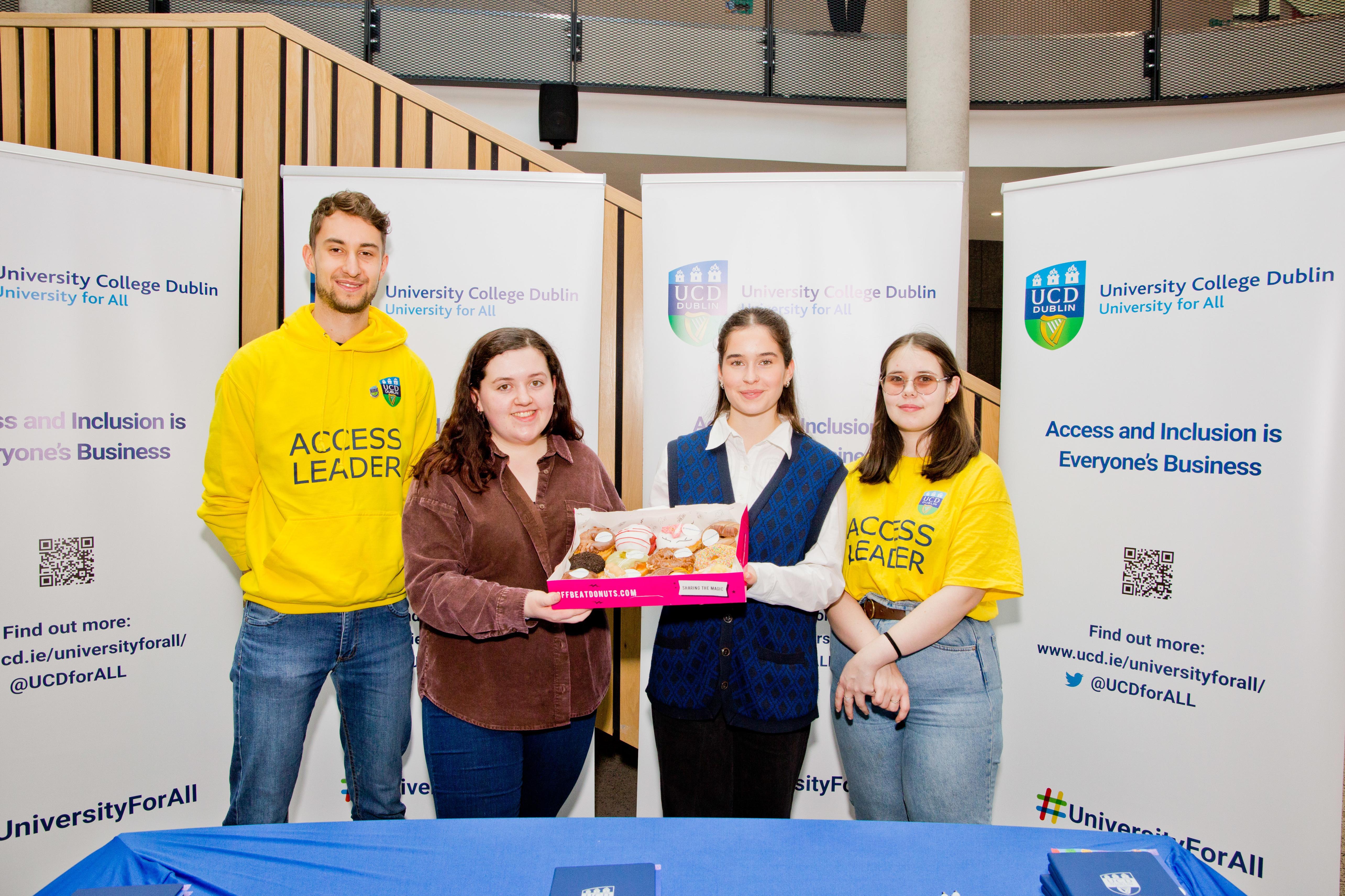 Four students standing in front of a blue table, the 2 students in the middle are holding a box of donuts. There are four white popup signs at the back.